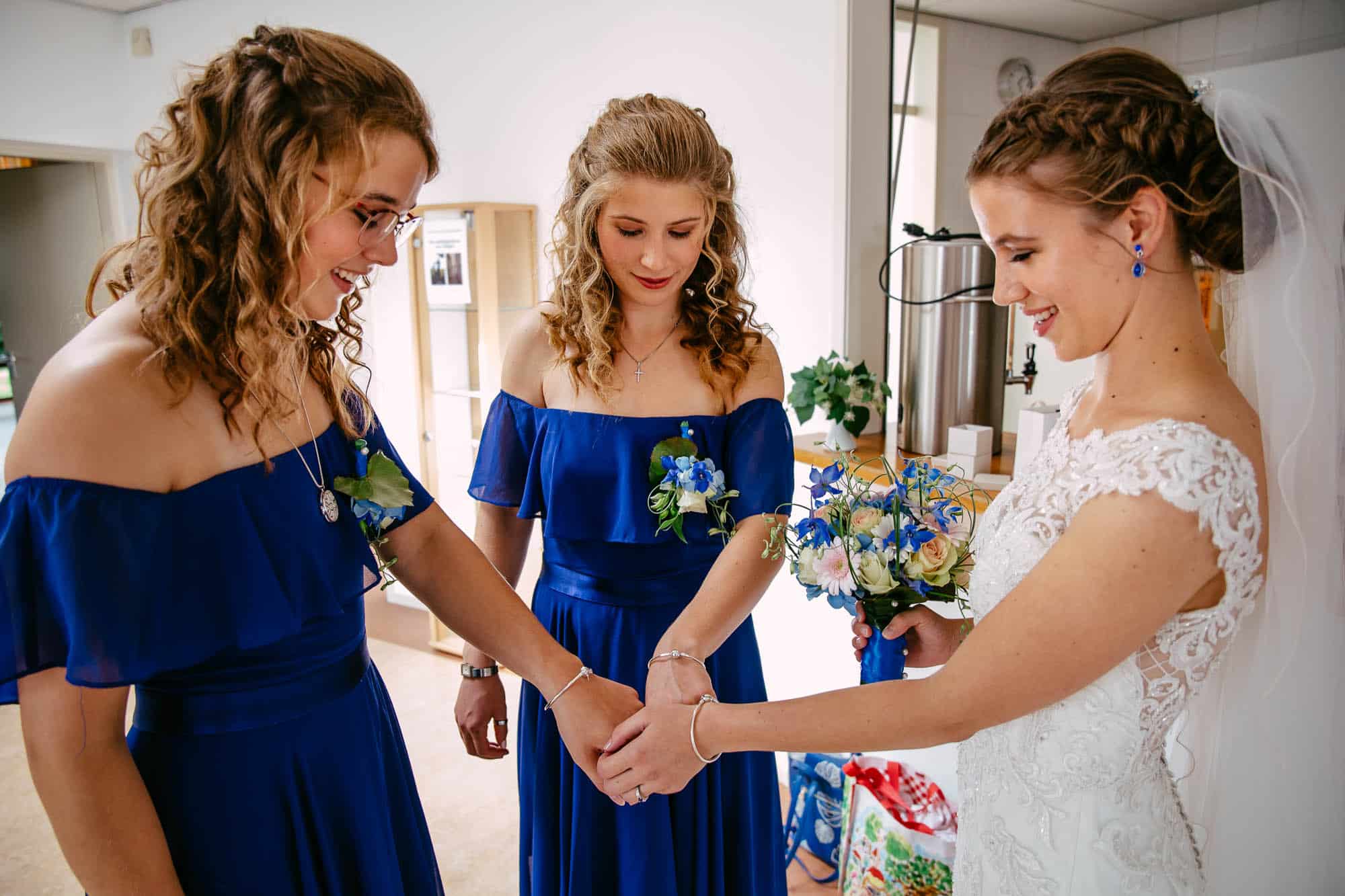 Drie bruidsmeisjes in een blauwe jurk, waarbij één ceremoniemeester elkaars hand vasthoudt.
