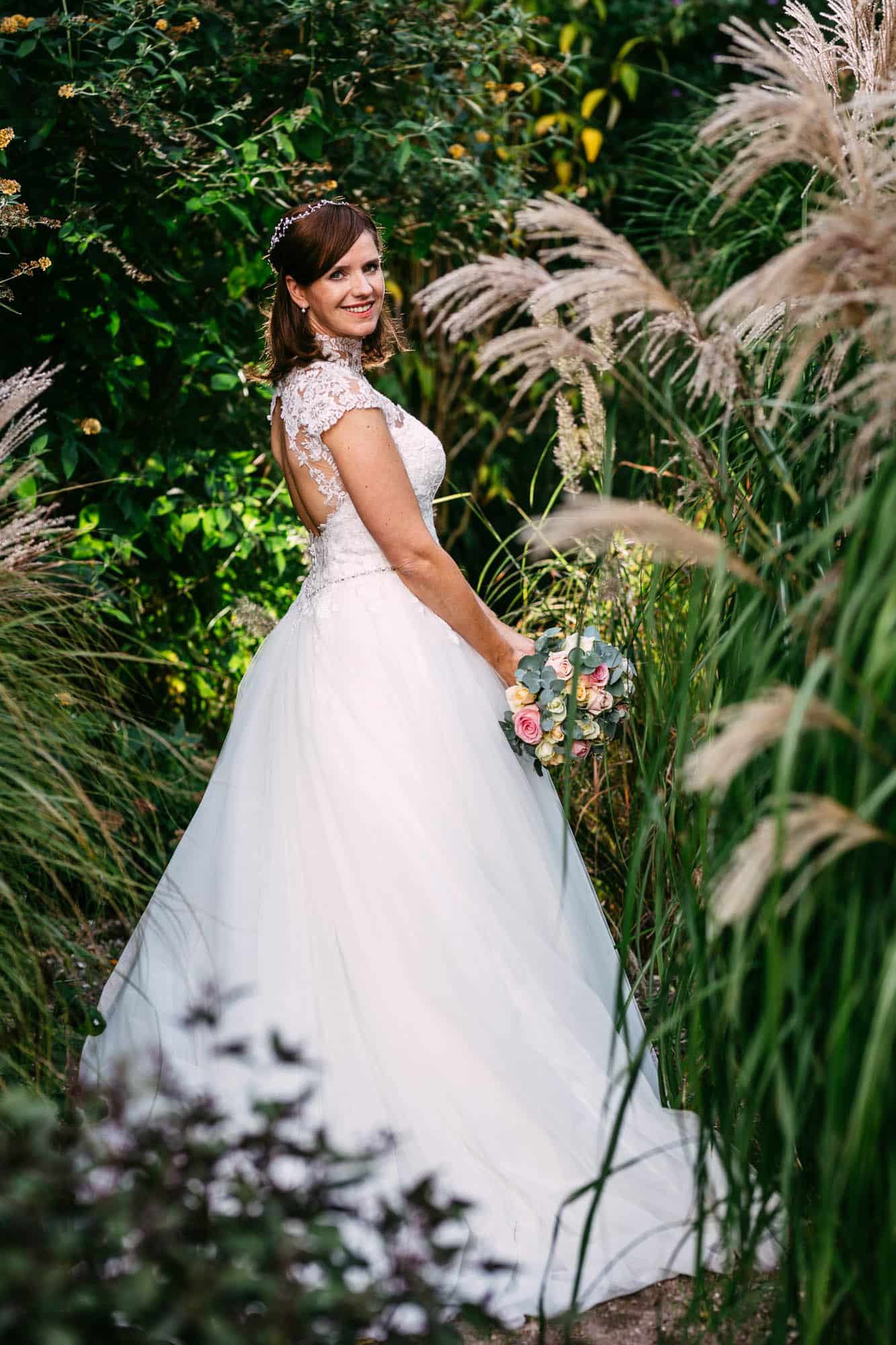 A bride in an A-line wedding dress, standing in a garden with tall reeds.