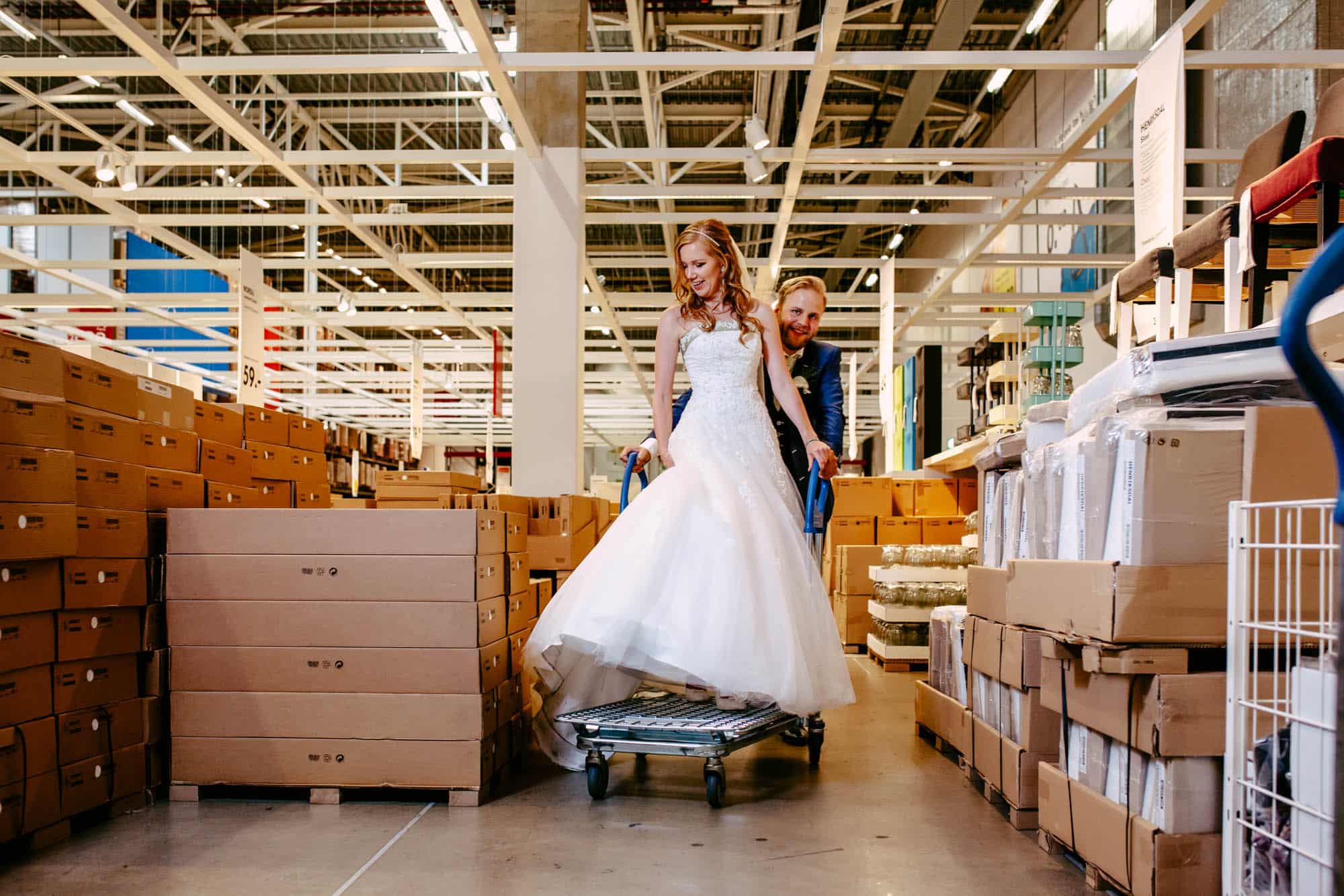 A bride and groom buying furniture in an IKEA warehouse, with the bride wearing a beautiful A-line wedding dress.