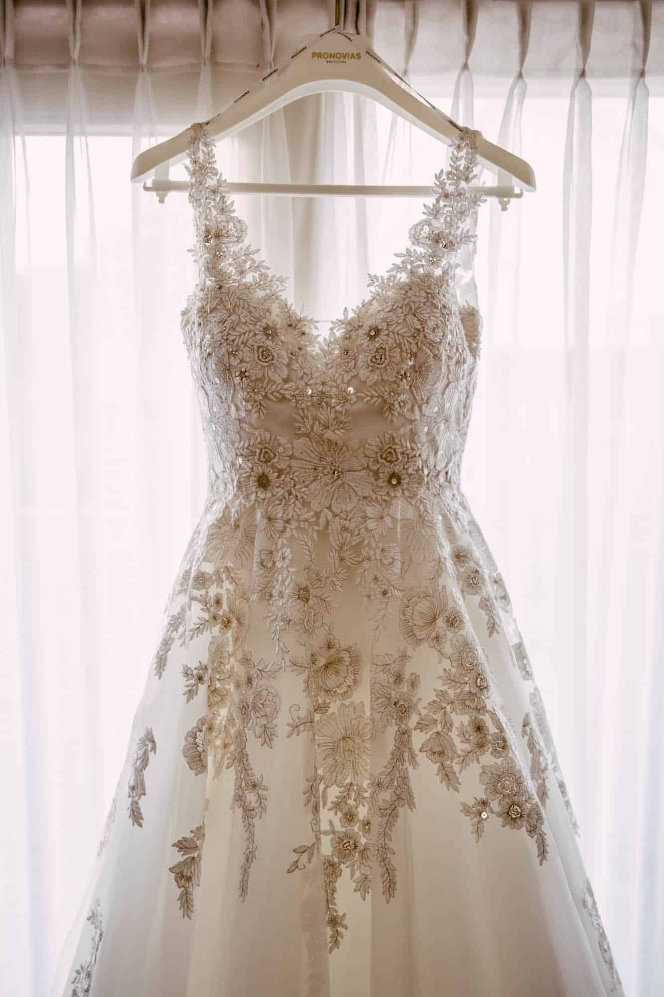 A wedding dress hangs from a hanger in front of a window.