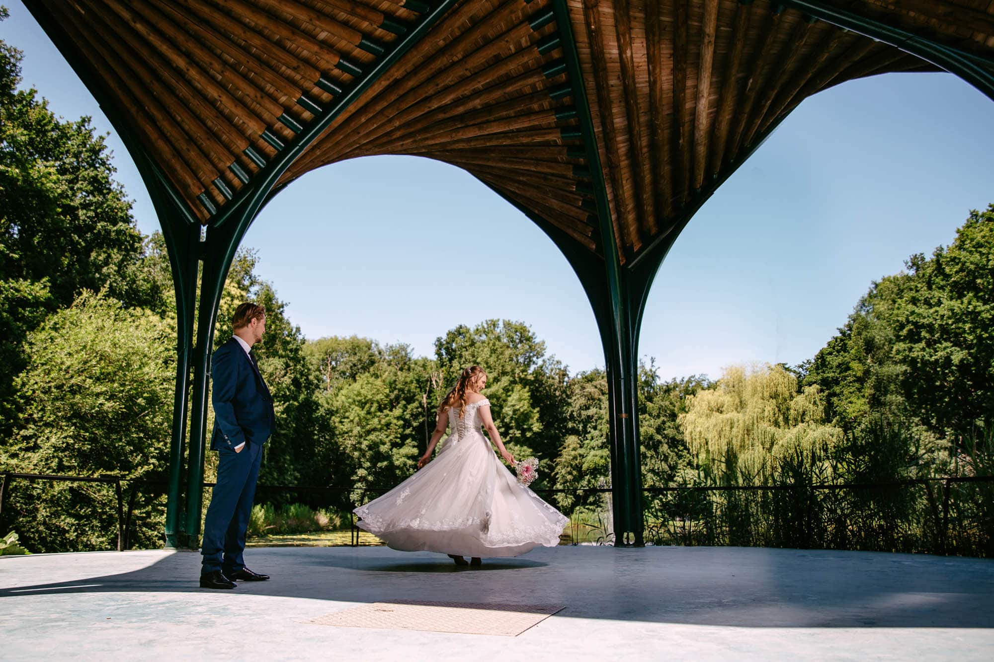 Een bruid en bruidegom staan onder een tuinhuisje, de bruid draagt een prachtige A-lijn trouwjurk.