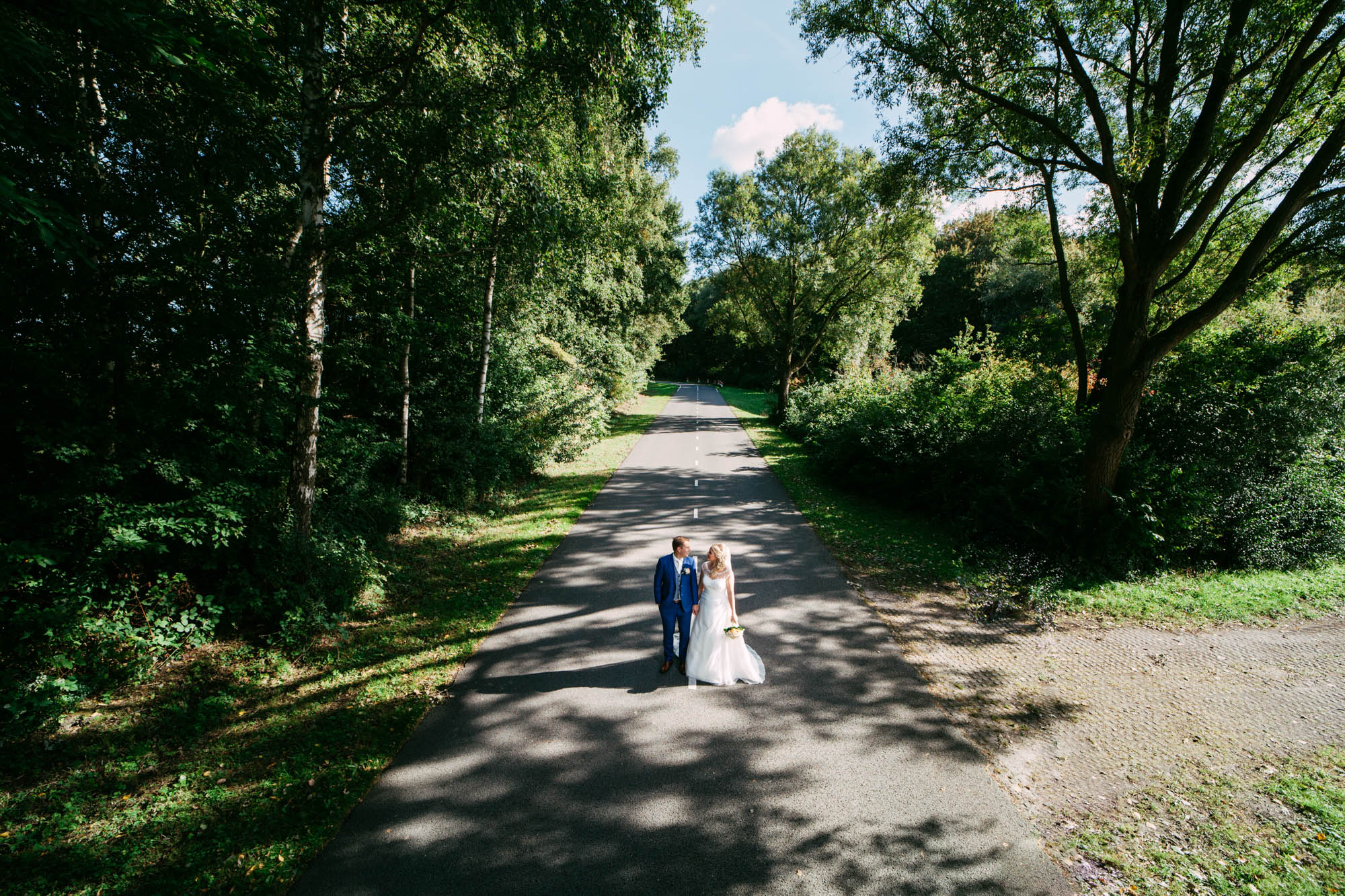 A bride in an A-line wedding dress and groom stand on a path in the forest.