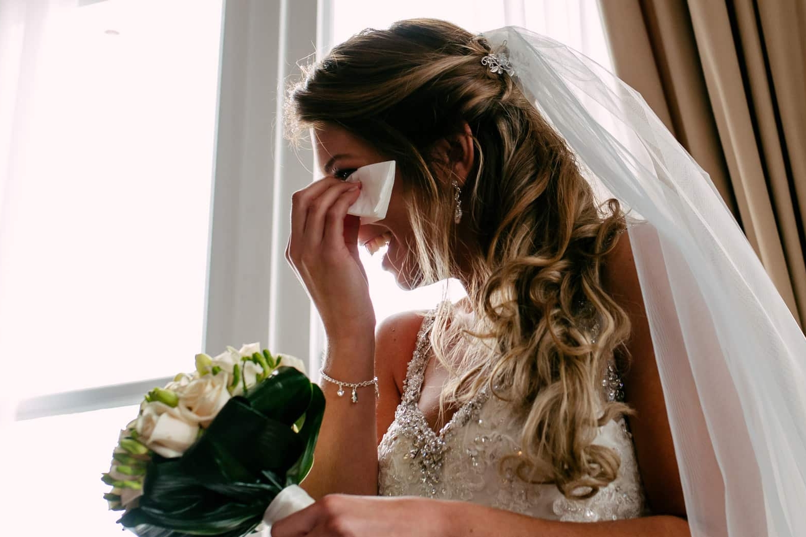 A bride in wedding dress wiping her eyes with a tissue during wedding vows.