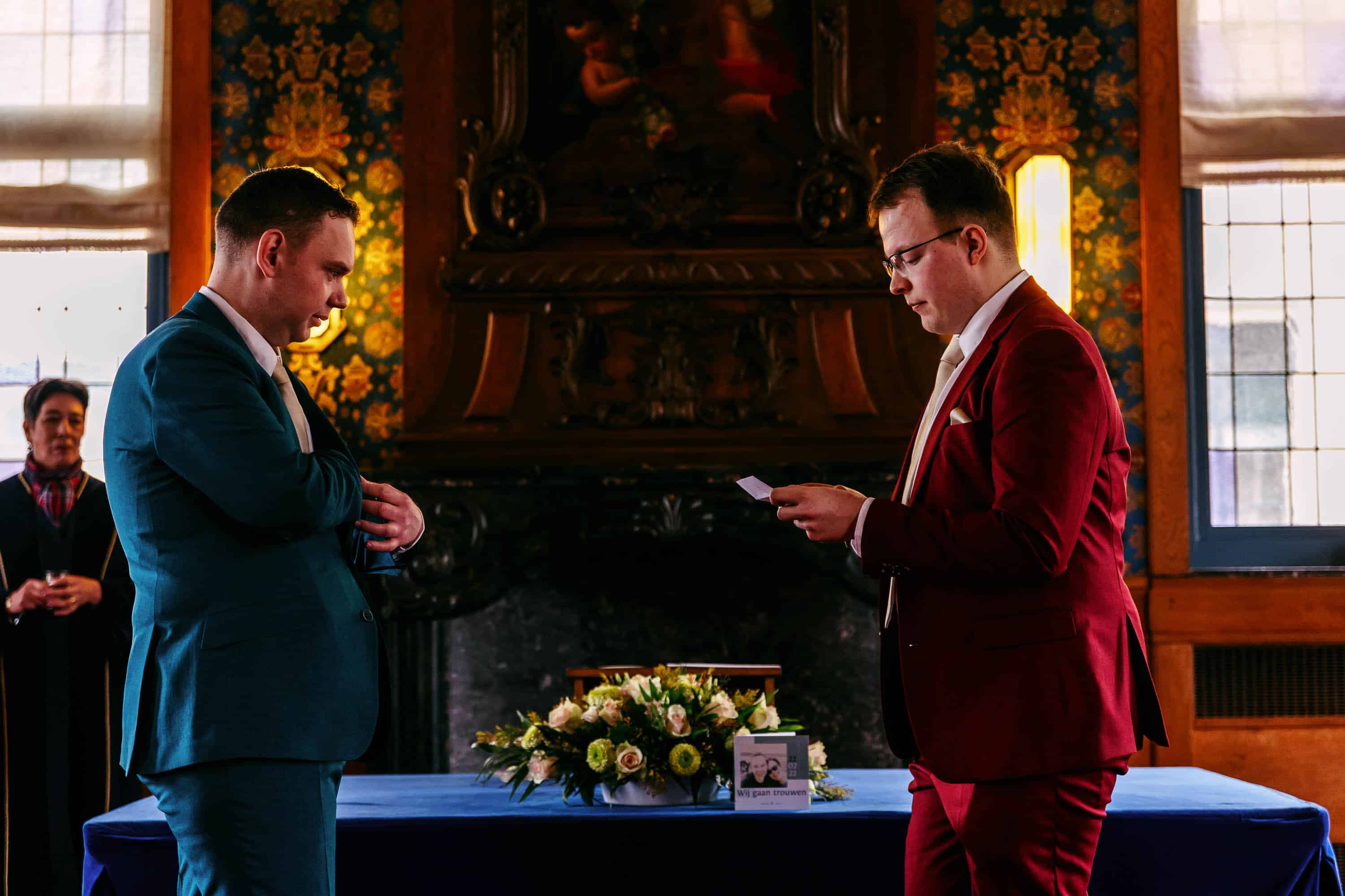 Two men in wedding-themed suits look at each other at a table.