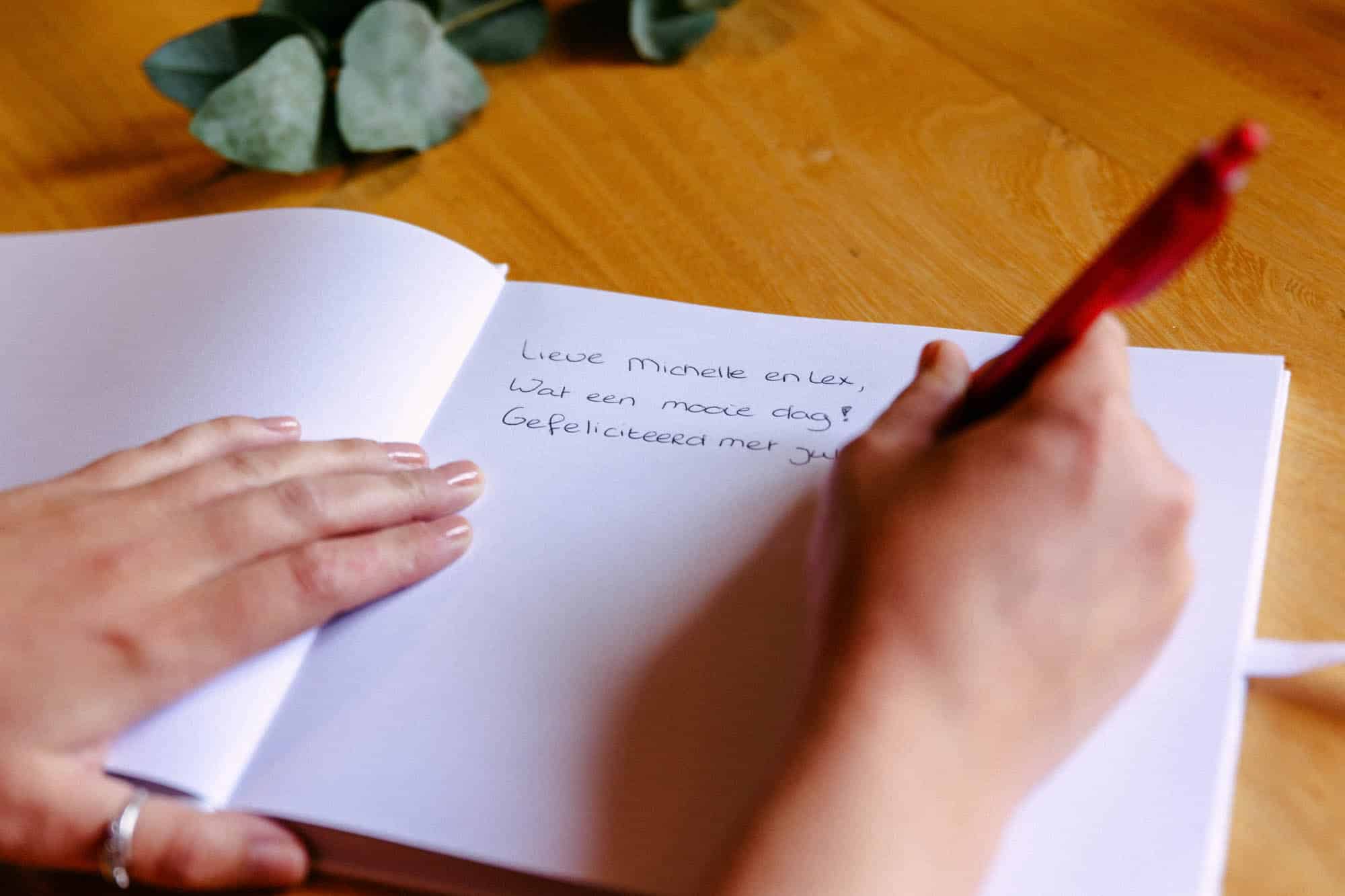 Een persoon die felicitaties voor een bruiloft opschrijft in een notitieboekje, met behulp van een rode pen.
