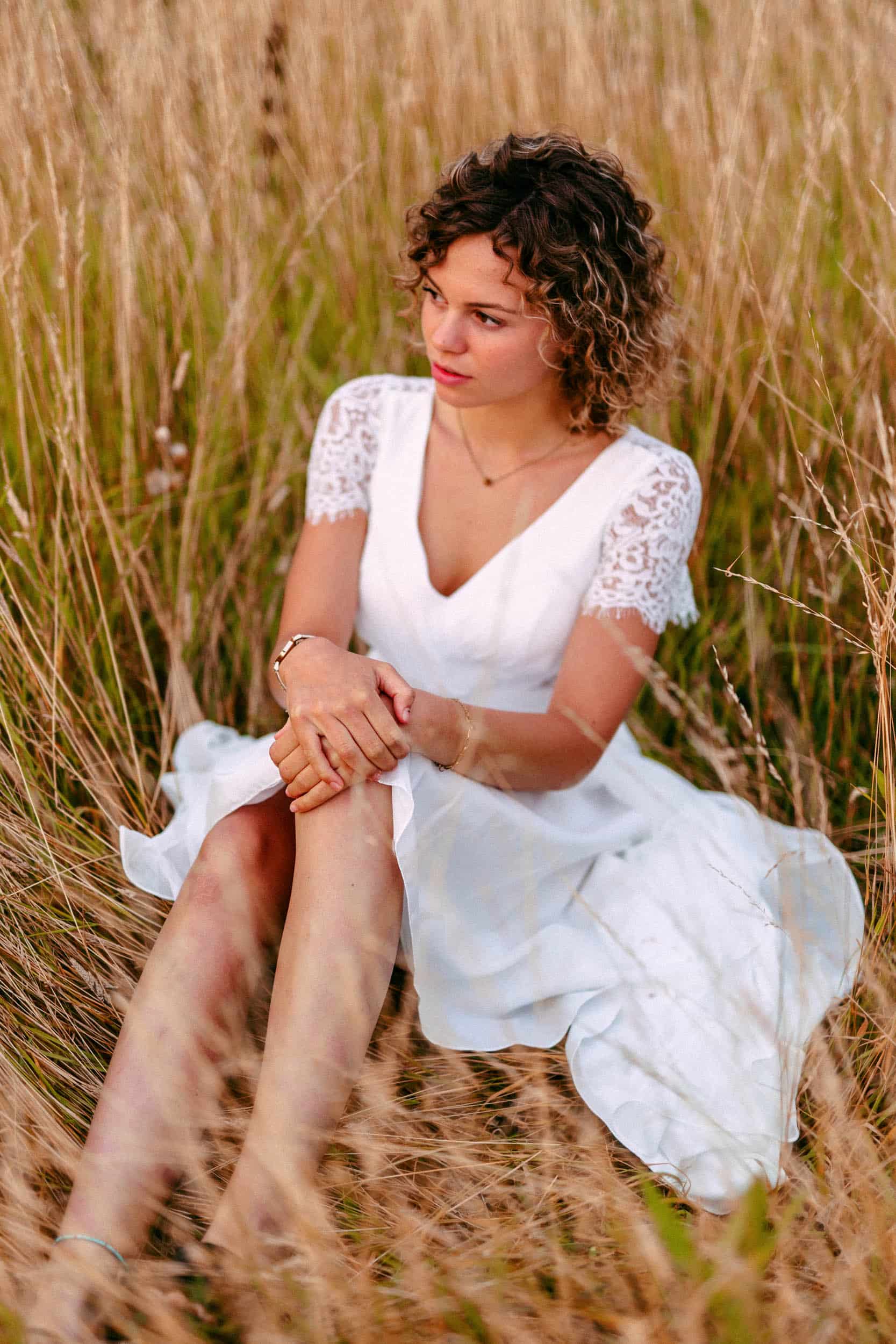 A woman in a white dress, starting to plan her wedding, while sitting in the tall grass.