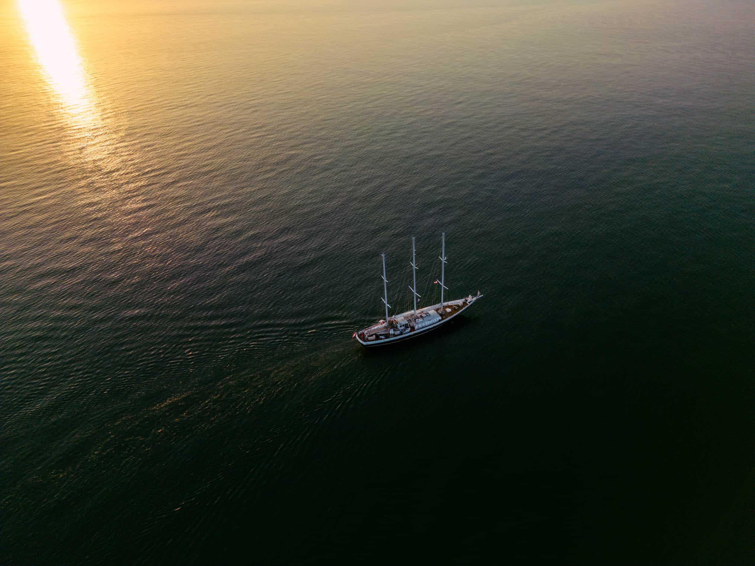 Een zeilboot glijdt gracieus door het schilderachtige oceaanwater bij zonsondergang, prachtig vastgelegd door een drone piloot huren.