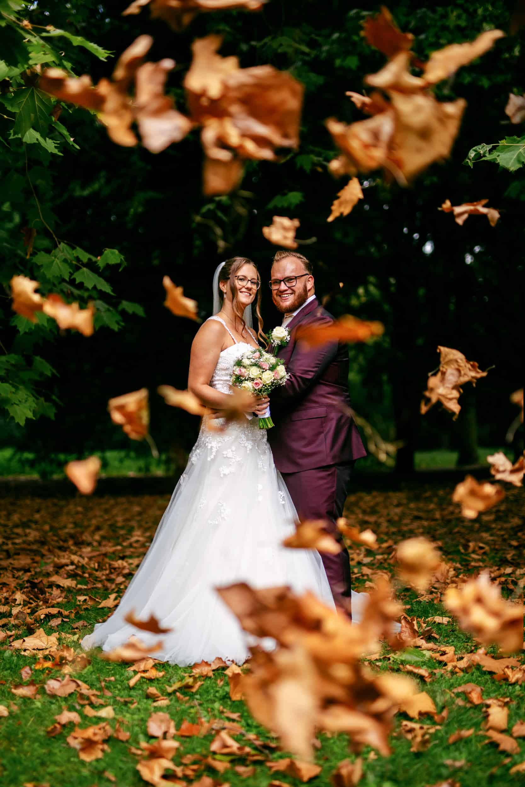 A bride and groom stand in a field with wedding-themed leaves falling around them.