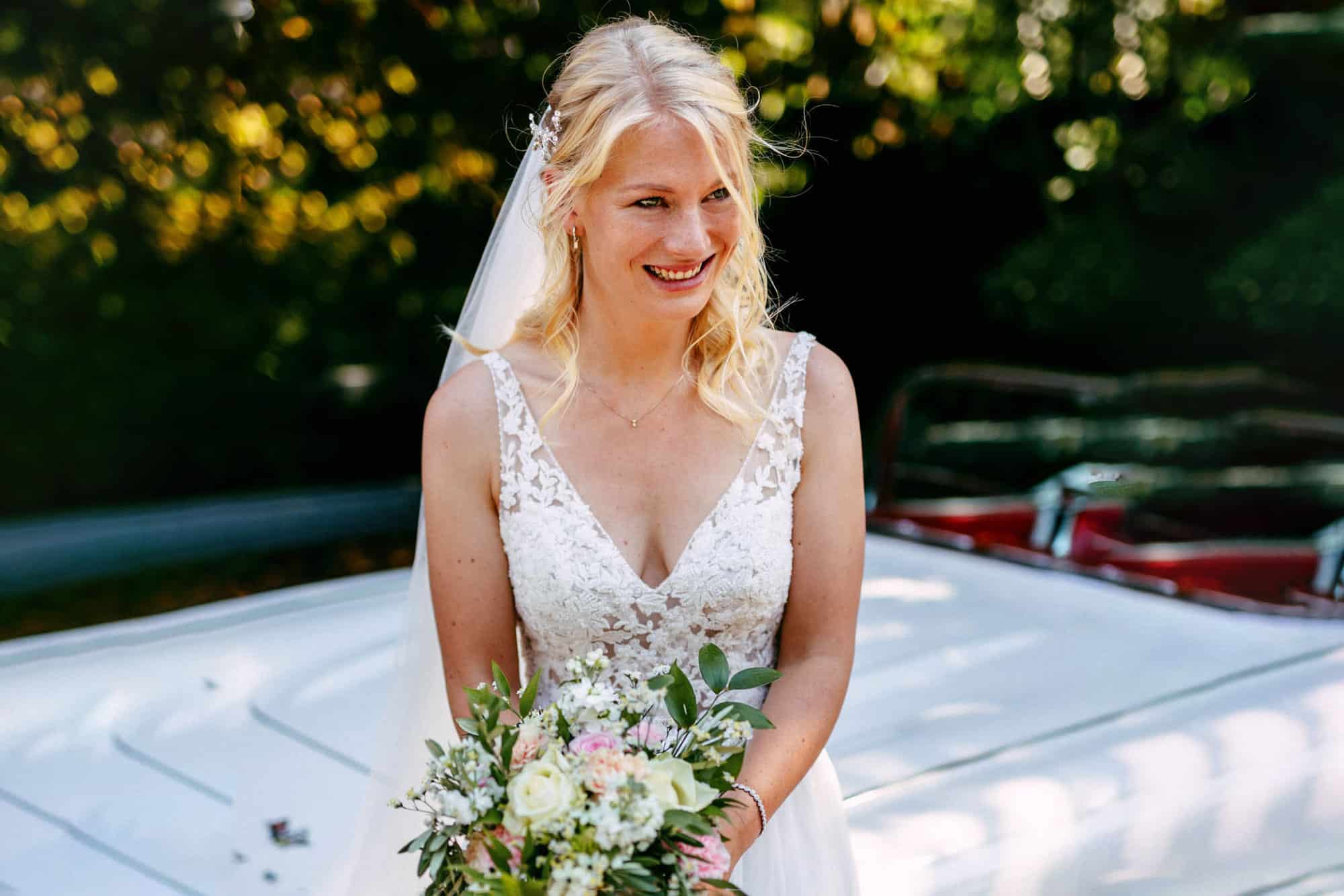 A bride in a wedding dress stands in front of a classic car.