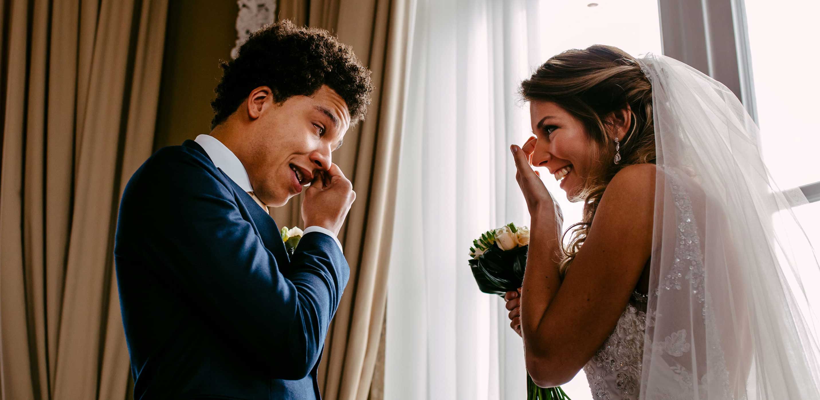 Een bruid en bruidegom die een luchtig moment beleven voor een raam, vastgelegd door een trouwfotograaf.