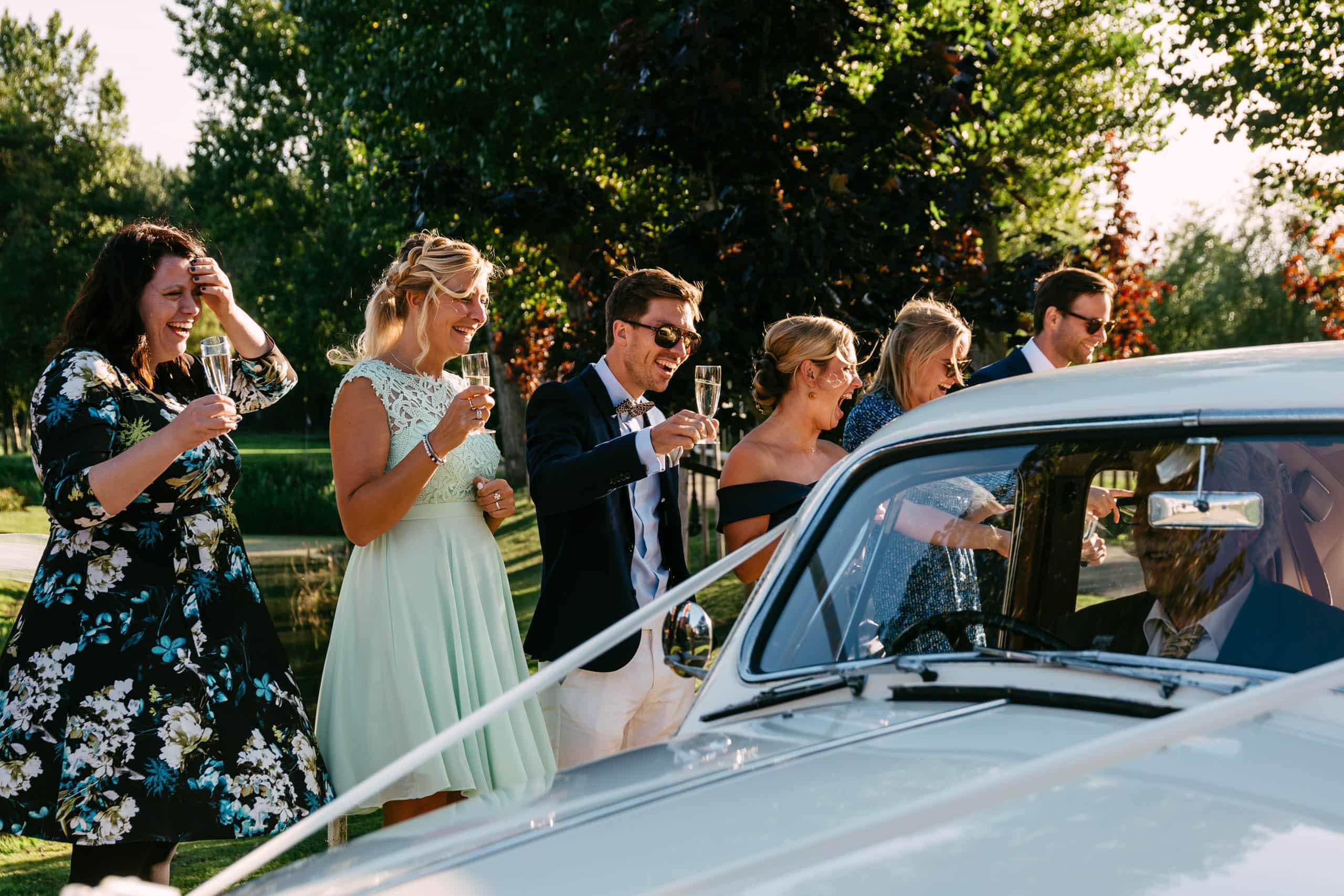 A group of bridesmaids and groom toast in front of a vintage car during their wedding party.