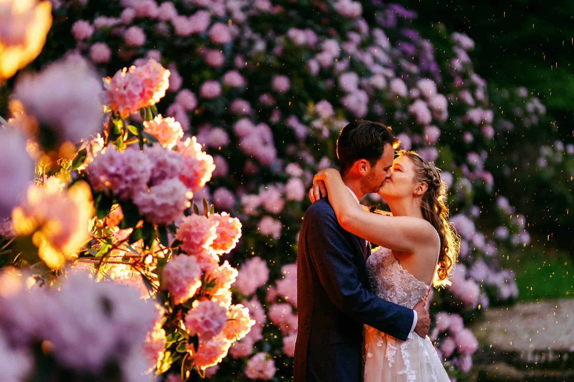 A bride and groom share a romantic kiss amid a vibrant floral display at their wedding, set against the backdrop of Rain at Your Wedding.