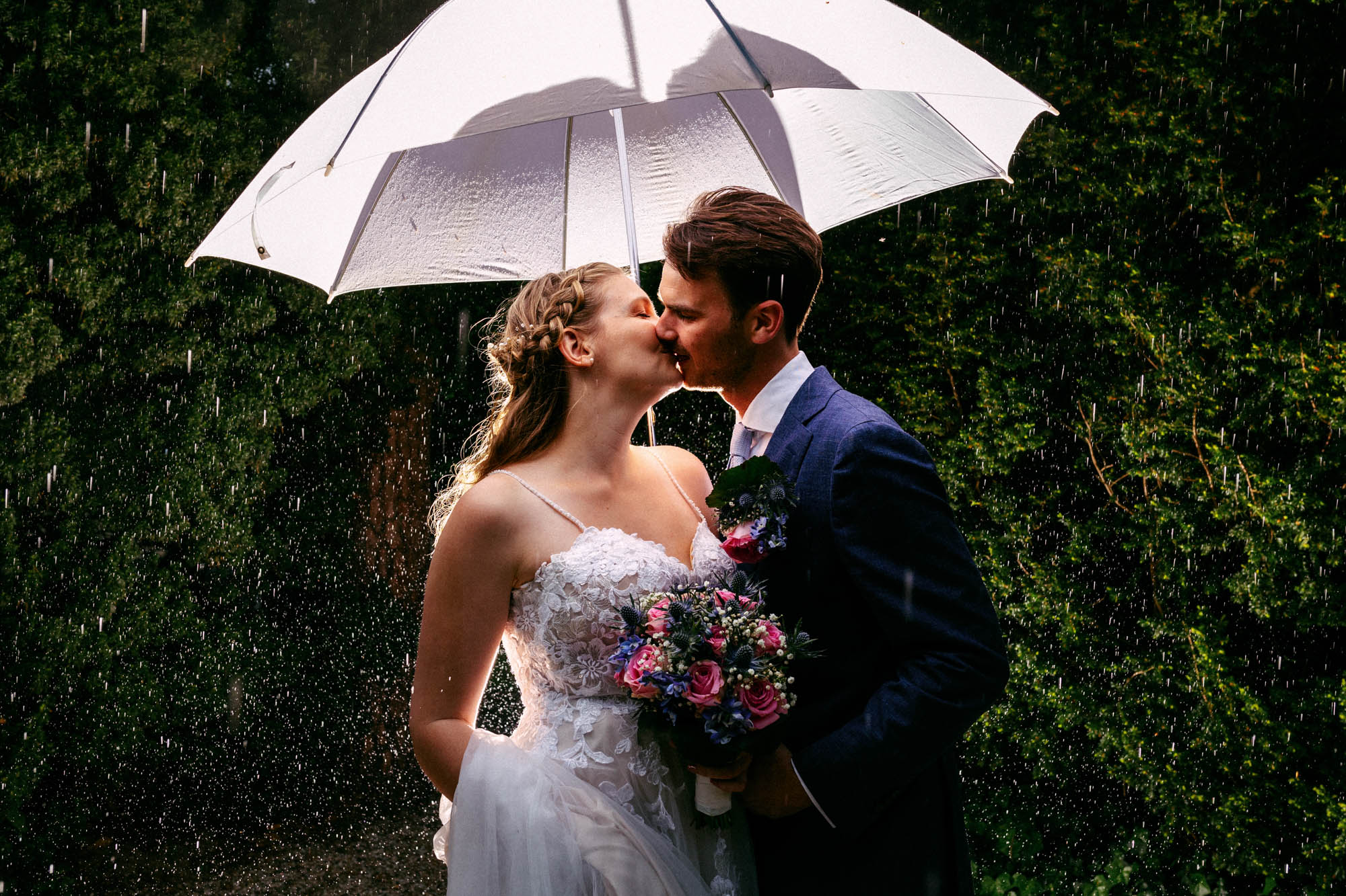 A bride and groom share a romantic kiss under an umbrella in the rain on their wedding day, experiencing the wonderful tradition of "Rain on your Wedding".