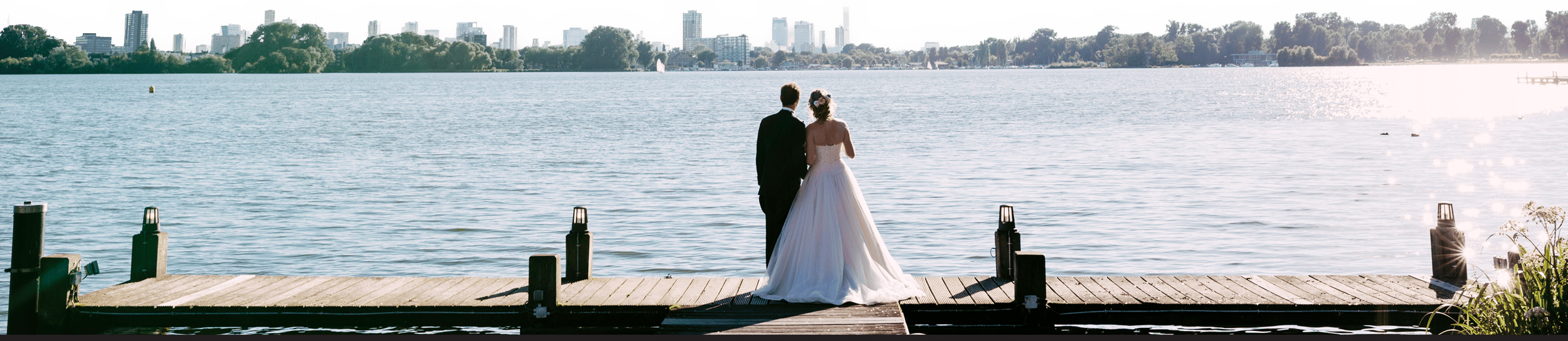 A bride and groom pose for their wedding photographer on a quay next to a picturesque lake.