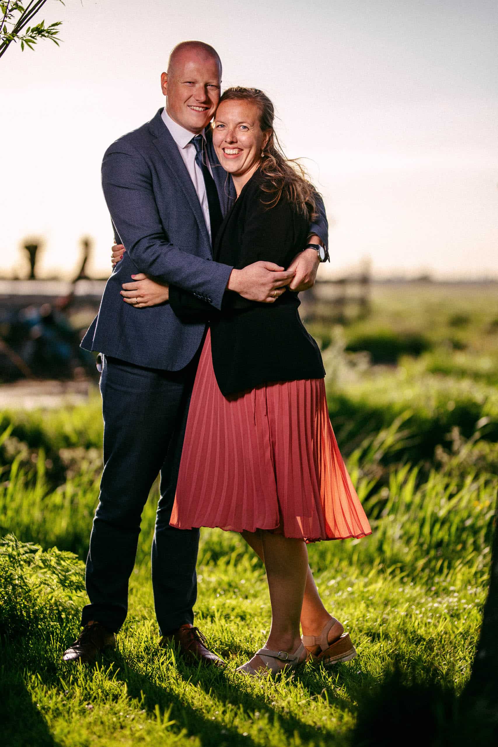 Een man en een vrouw die elkaar omhelzen in een veld bij zonsondergang, gekleed in Tenue de ville-kledij.