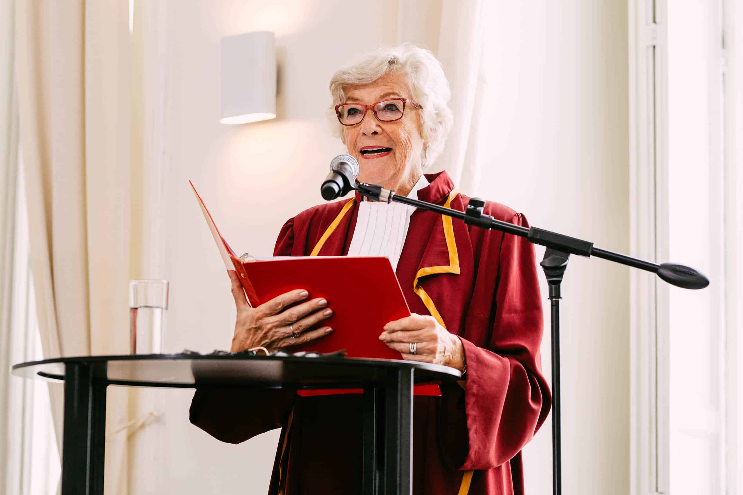 Een oudere vrouw in een rood gewaad staat op een podium tijdens een ondertrouwceremonie.