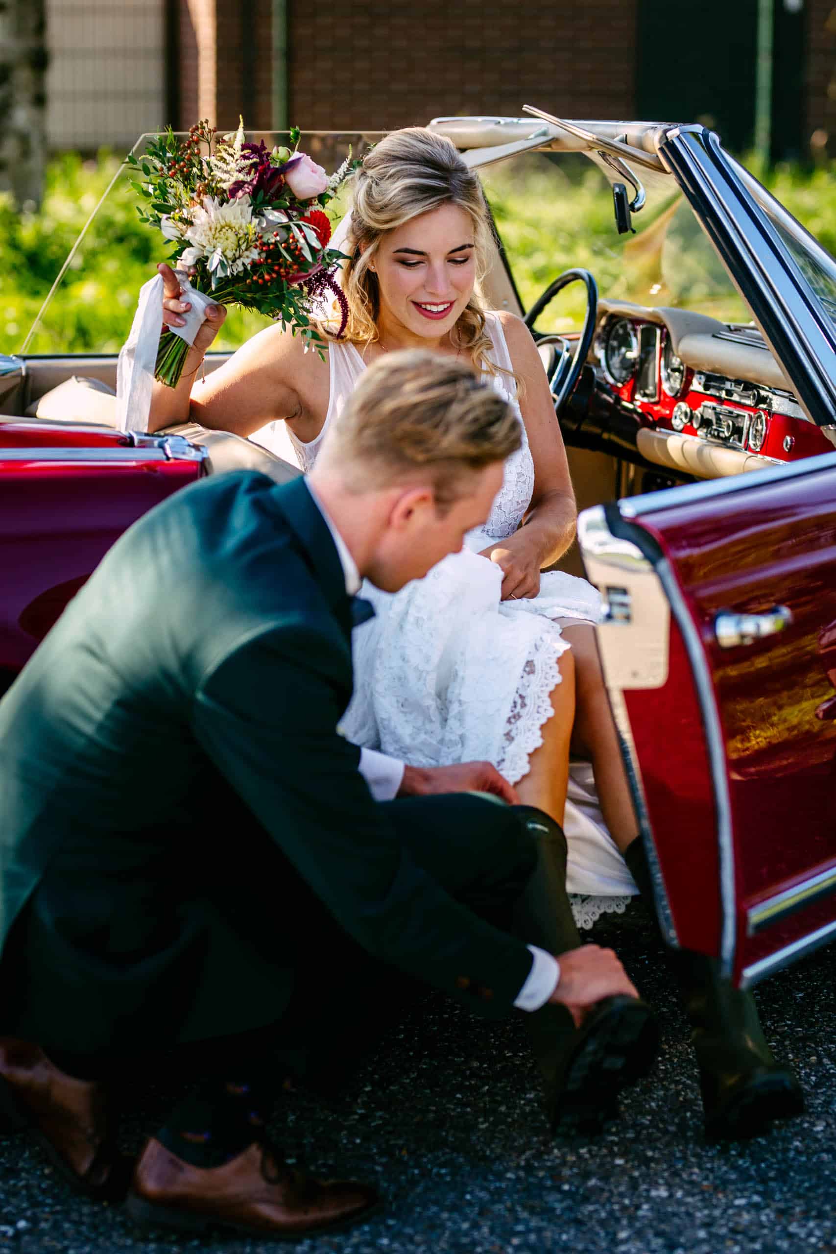 Een bruid en bruidegom zitten vrolijk in een rode cabriolet terwijl ze comfortabele bruidsschoenen dragen.
