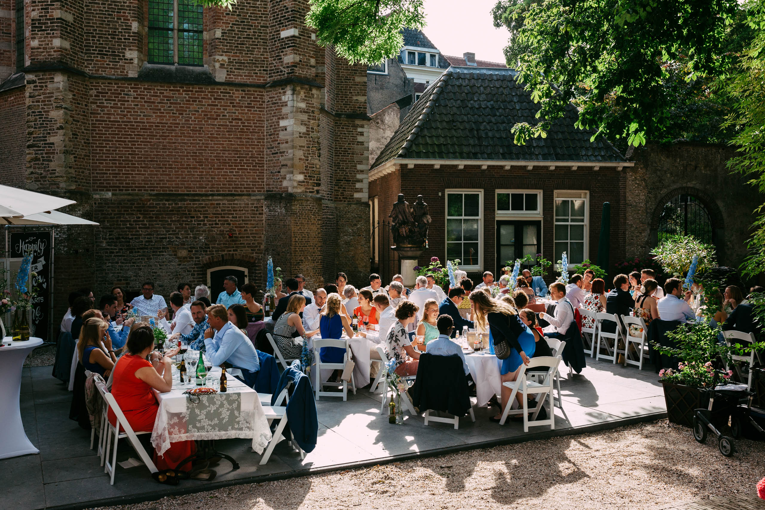 Een grote groep mensen zit aan tafels op een binnenplaats en viert een vreugdevolle bruiloft.