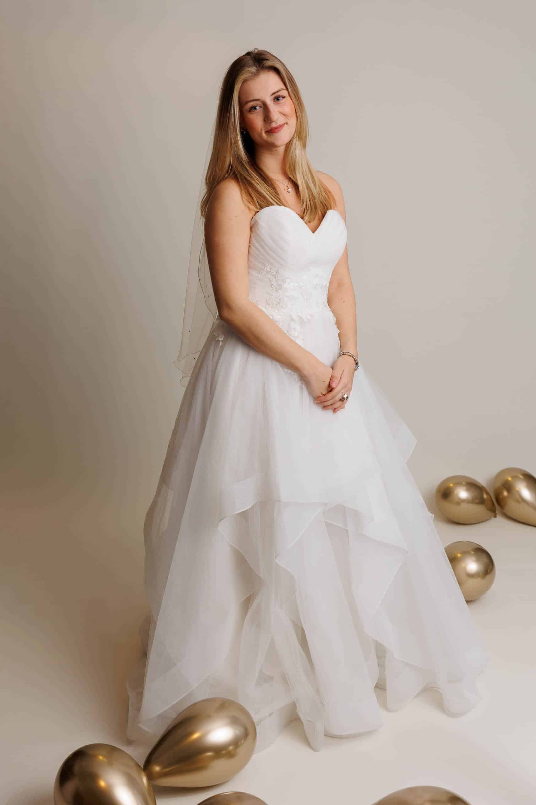 A bride in a wedding dress poses in front of balloons while trying on wedding dresses for fun.