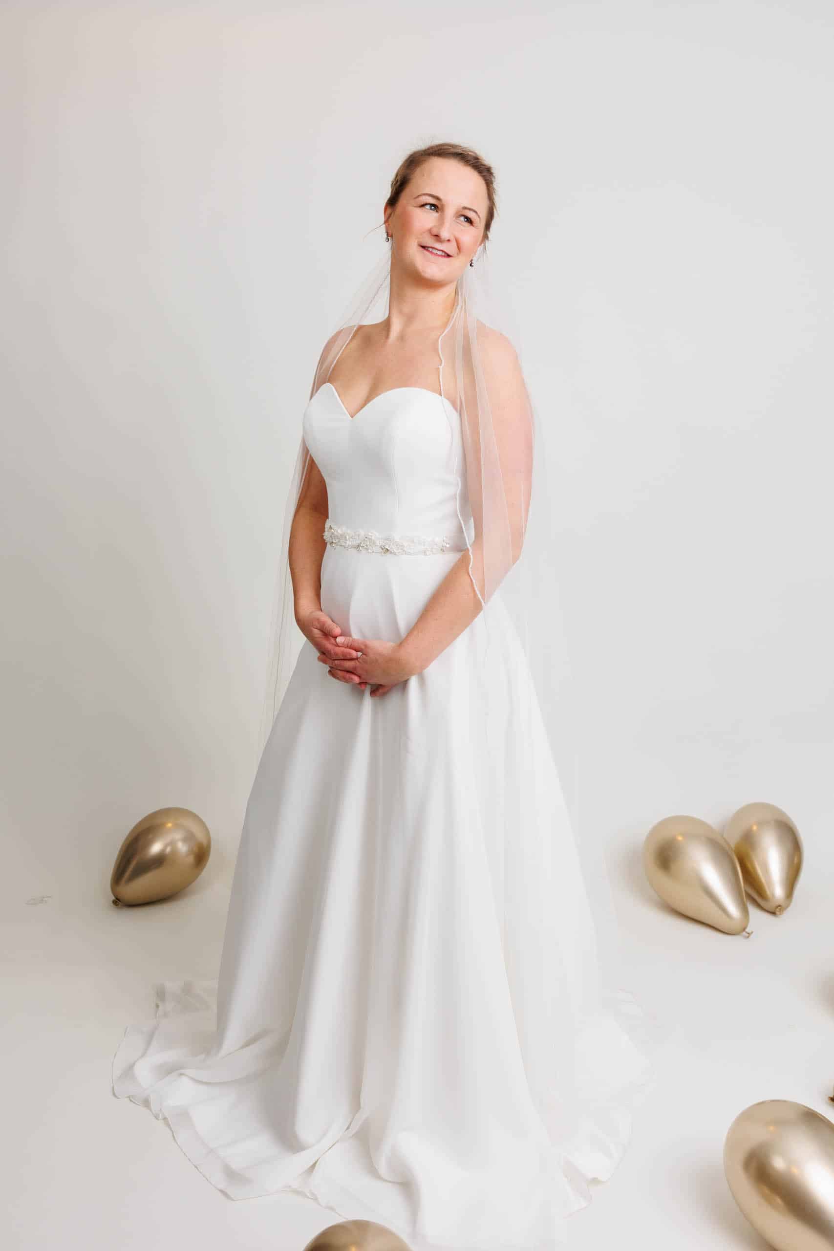 A bride wearing a white wedding dress poses in front of gold balloons while trying on wedding dresses for fun.