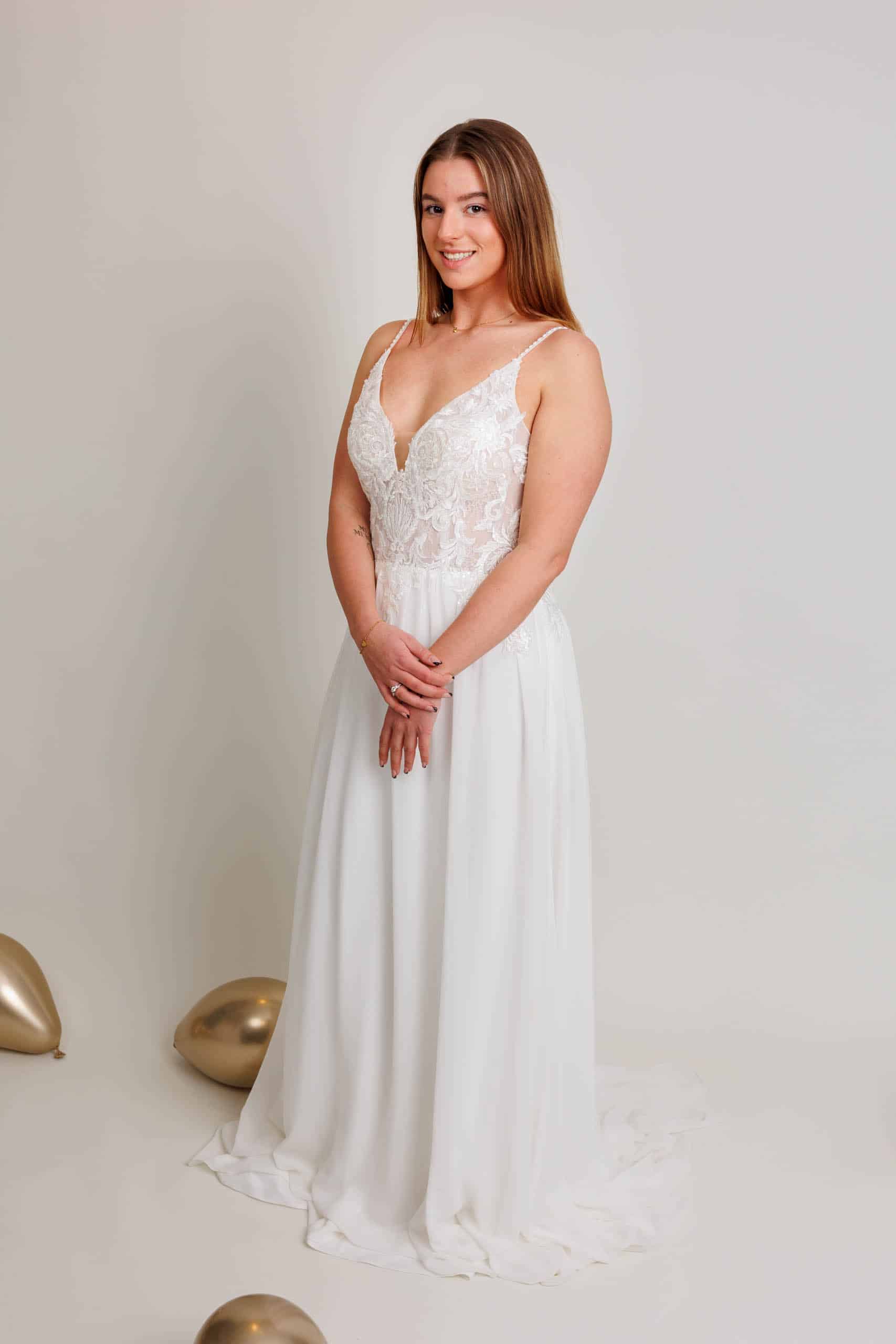A woman in a white wedding dress poses with gold balloons while trying on wedding dresses.