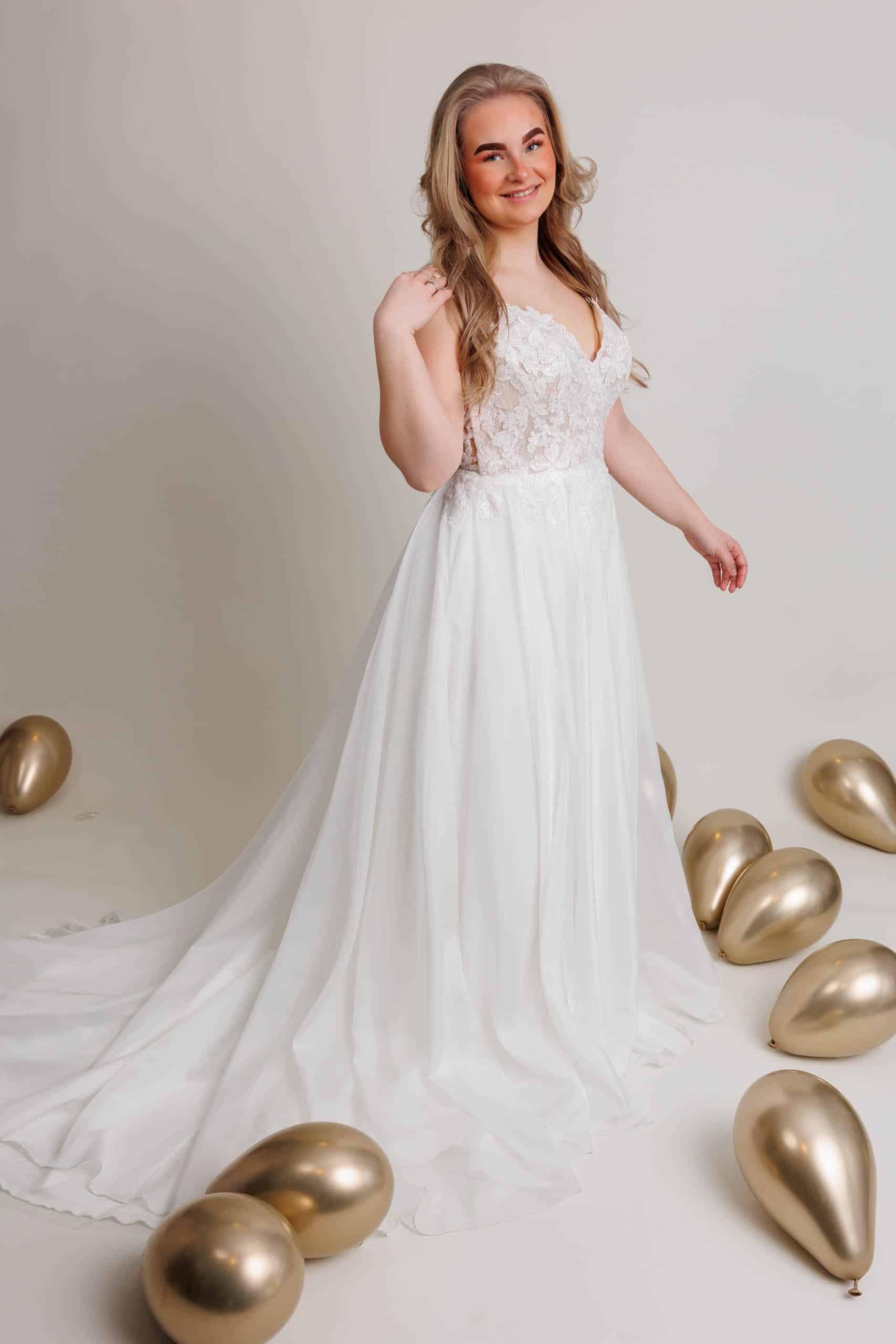 A woman in a white wedding dress poses with gold balloons while trying on wedding dresses for fun.