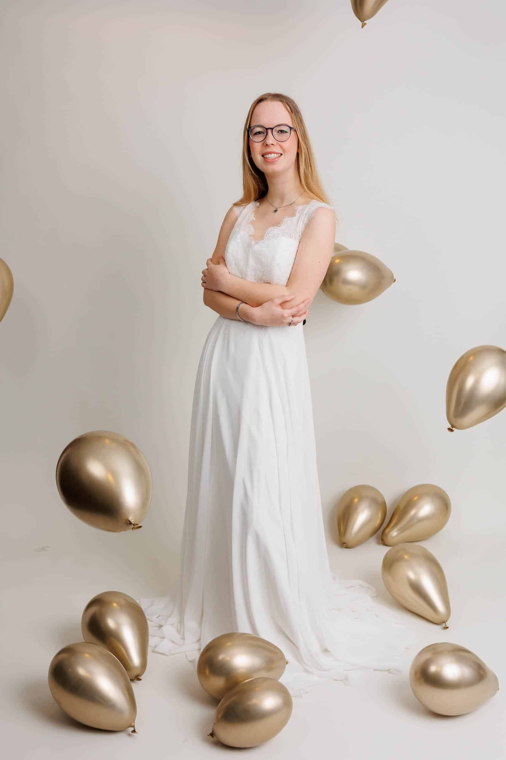 A woman in a wedding dress poses in front of gold balloons while trying on wedding dresses for fun.