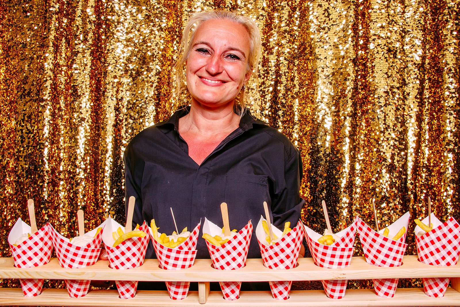 A woman standing in front of a gold background at a photobooth, holding food in cones.