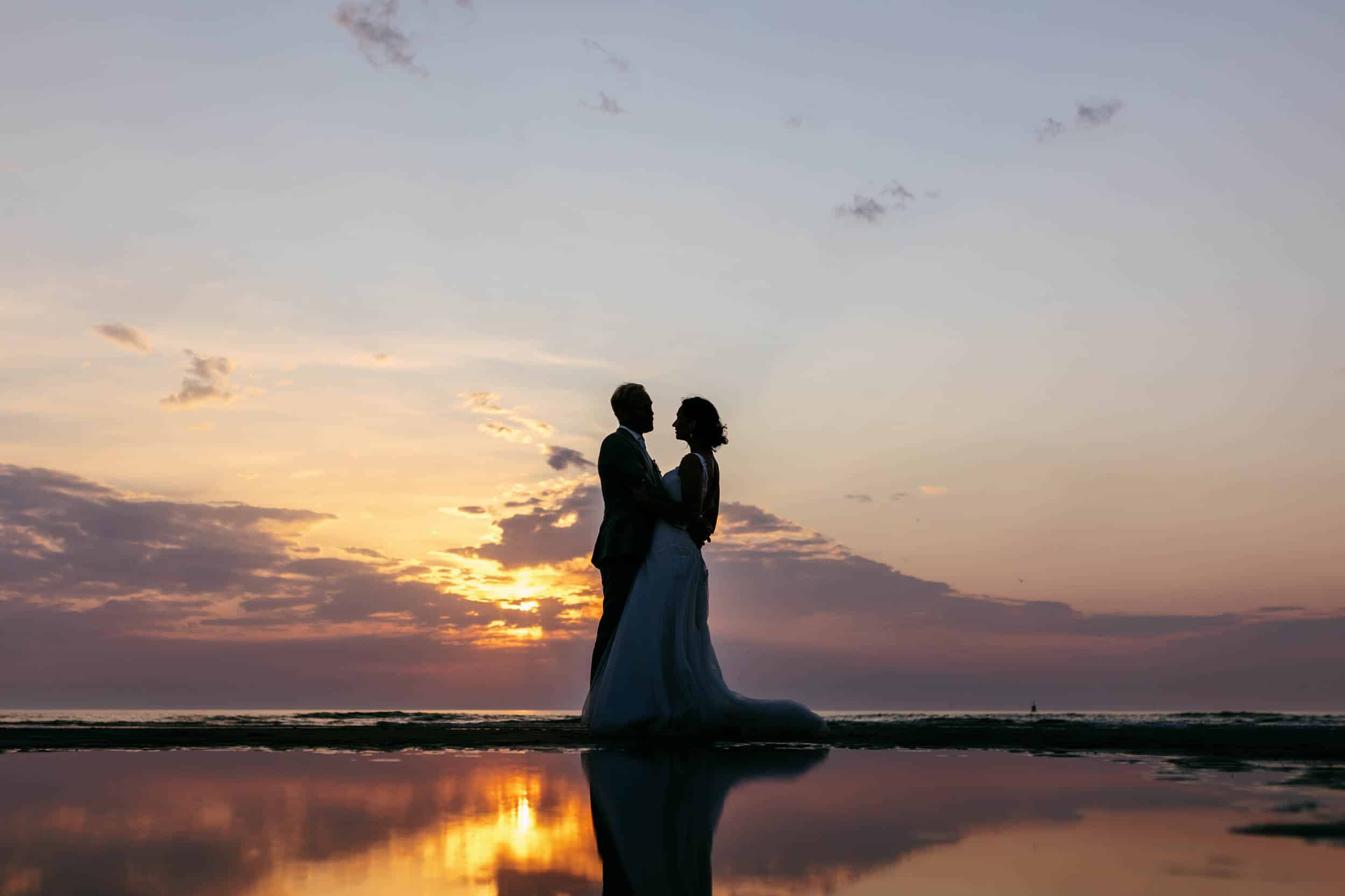 A wedding couple stand in the water on the beach at sunset.