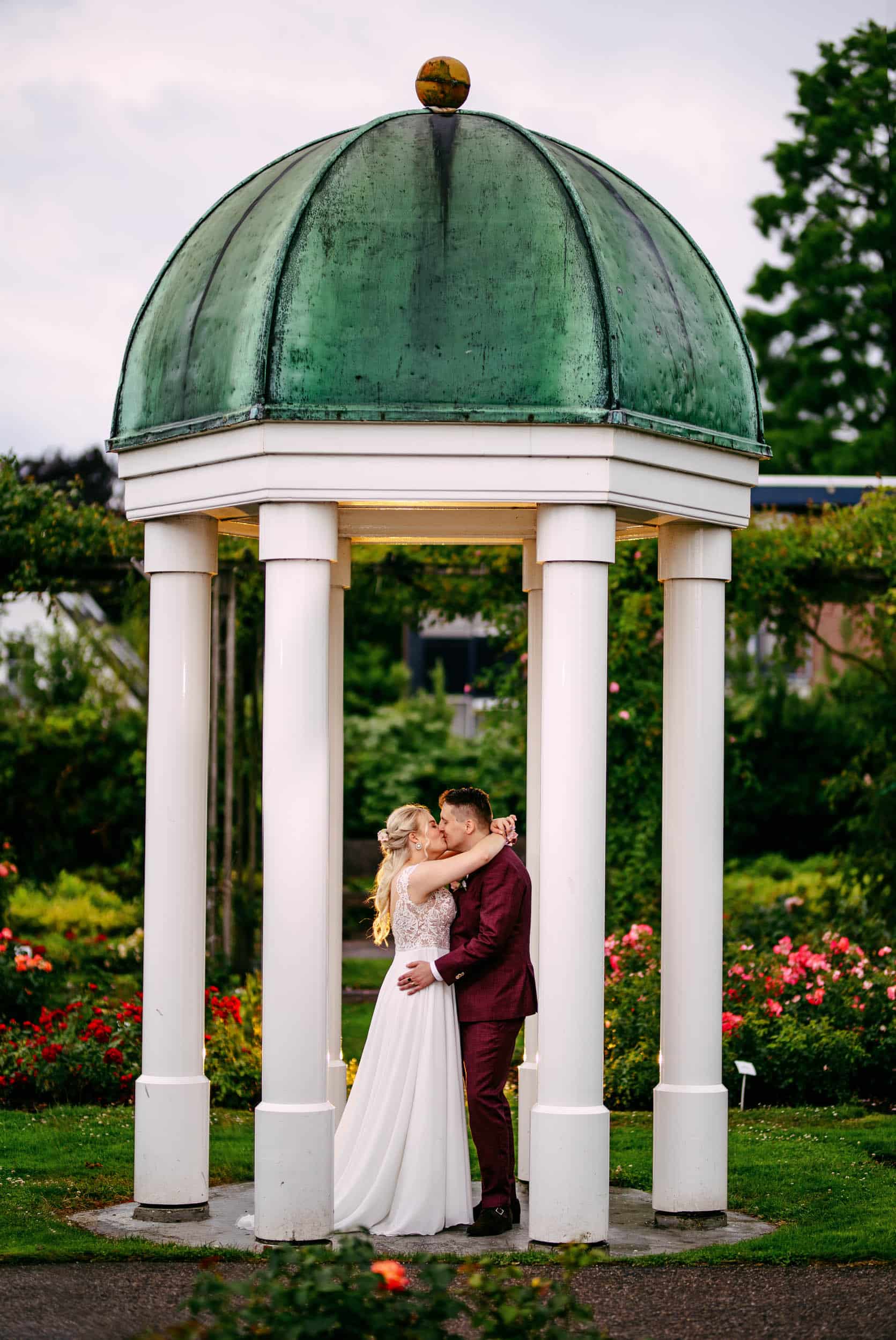 A bride and groom share a romantic kiss under an arbour in a beautifully landscaped garden, adding a historical element to their wedding celebration.