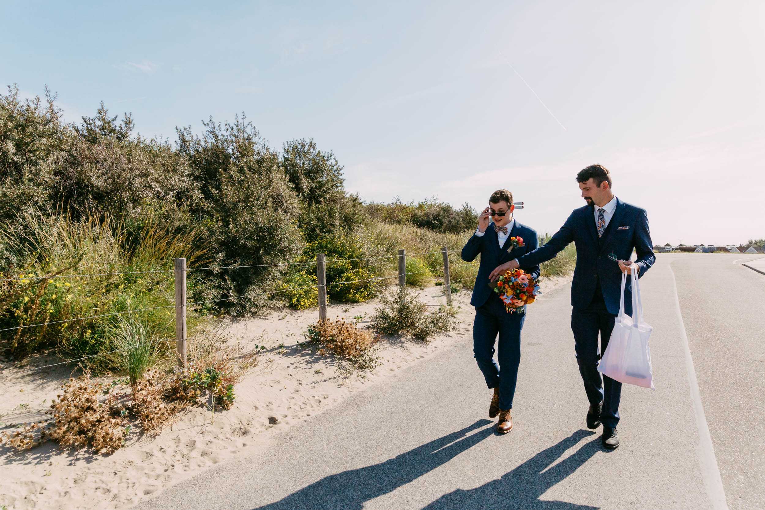 Two men in suits walk along a beach road.