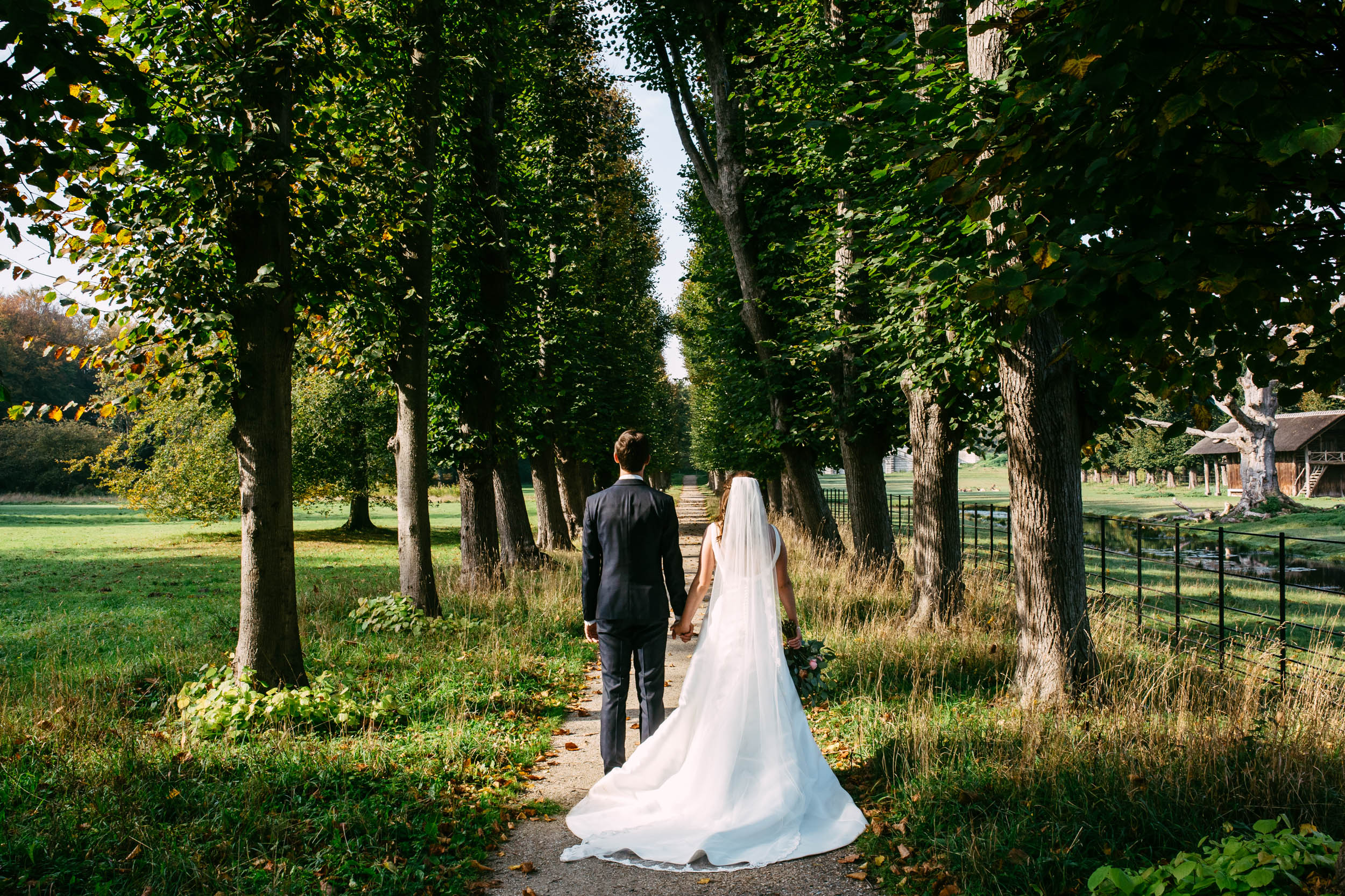 A bride and groom walk along a path lined with trees.