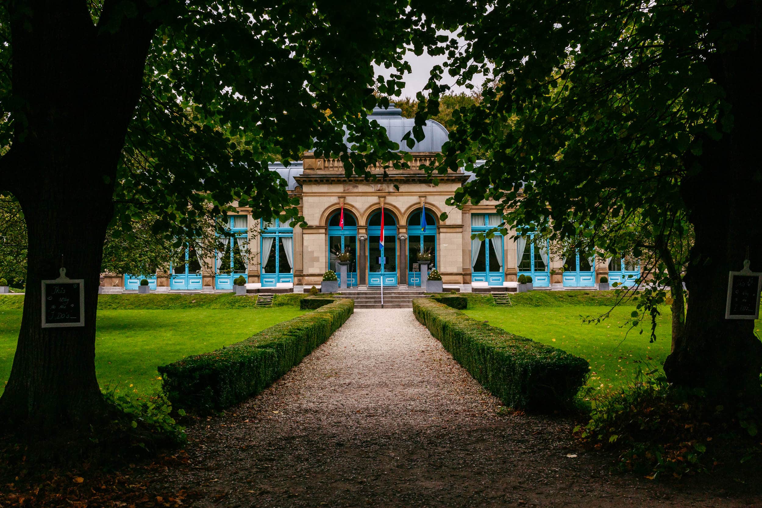 A path leading to a large building with blue doors called Orangerie Elswout.