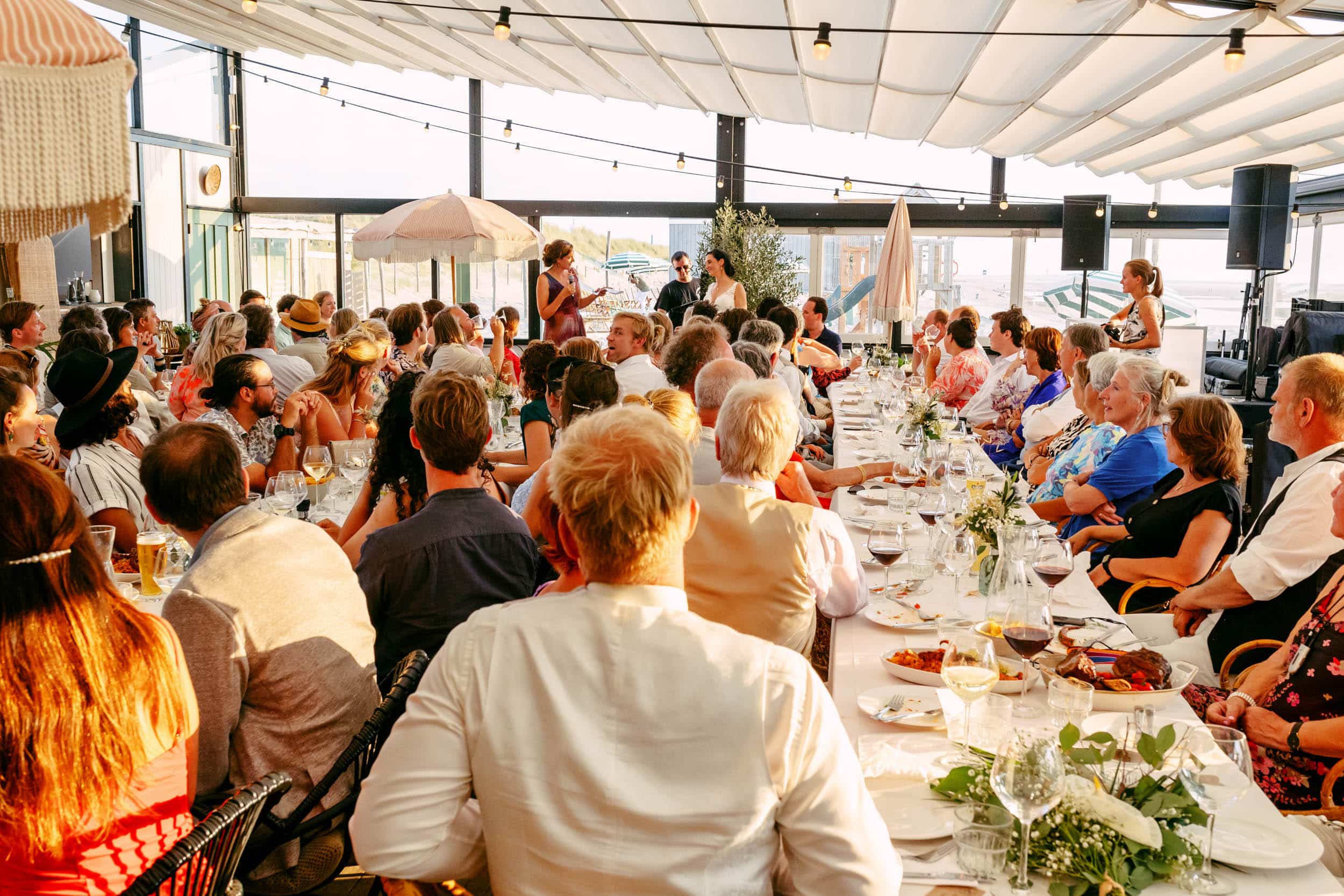 Een grote groep mensen zit aan tafels tijdens een bruiloftsreceptie in een strandclub.