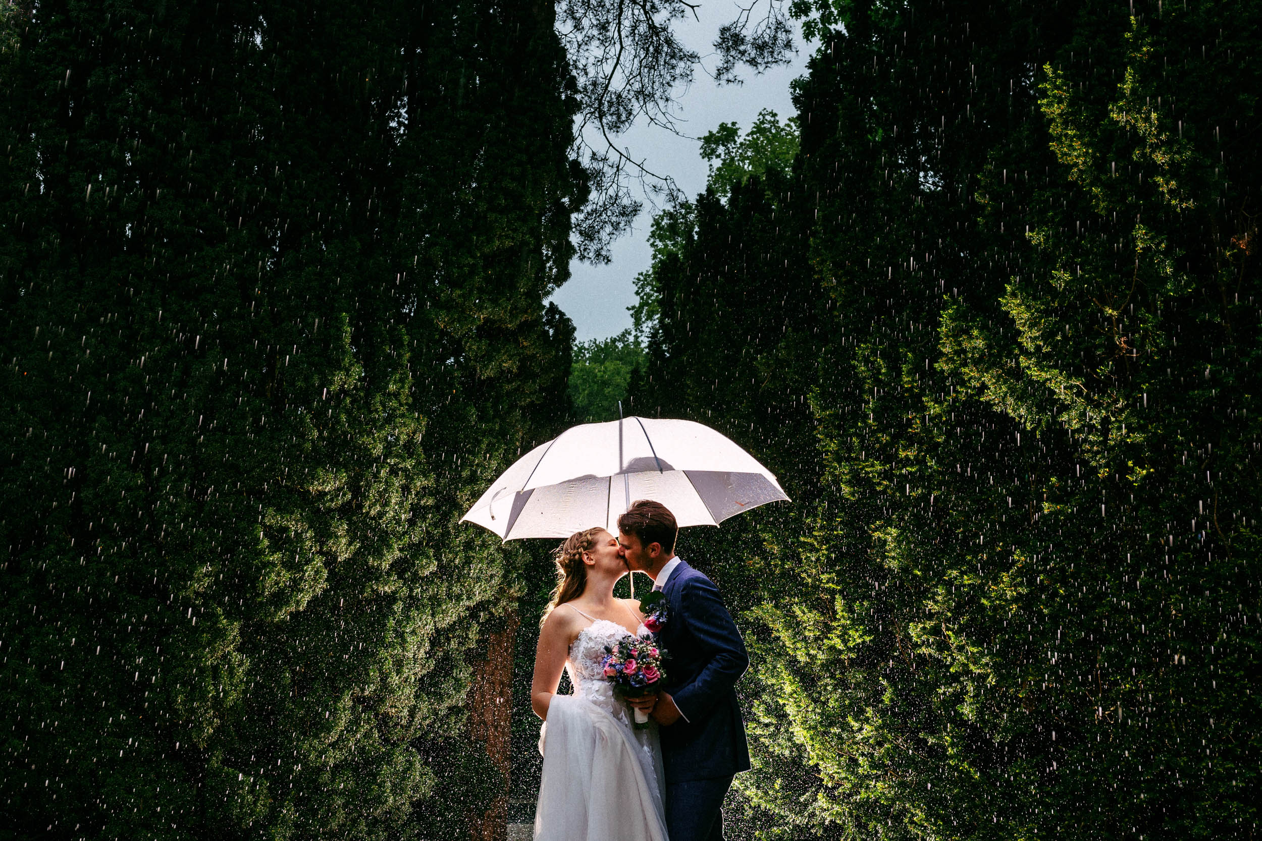 Een bruid en bruidegom kussen onder een paraplu in de regen.