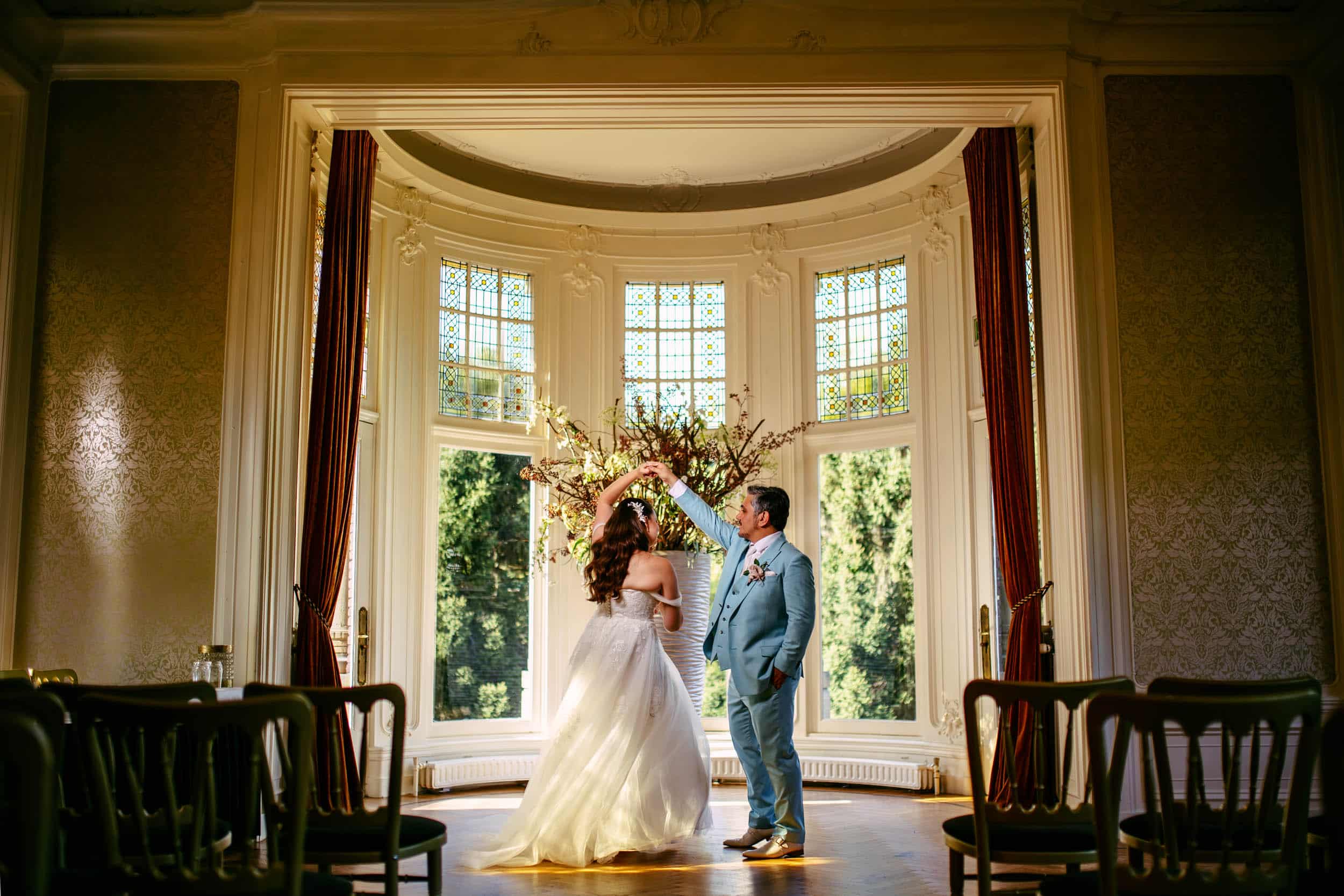 A bride and groom stand in front of a large window.