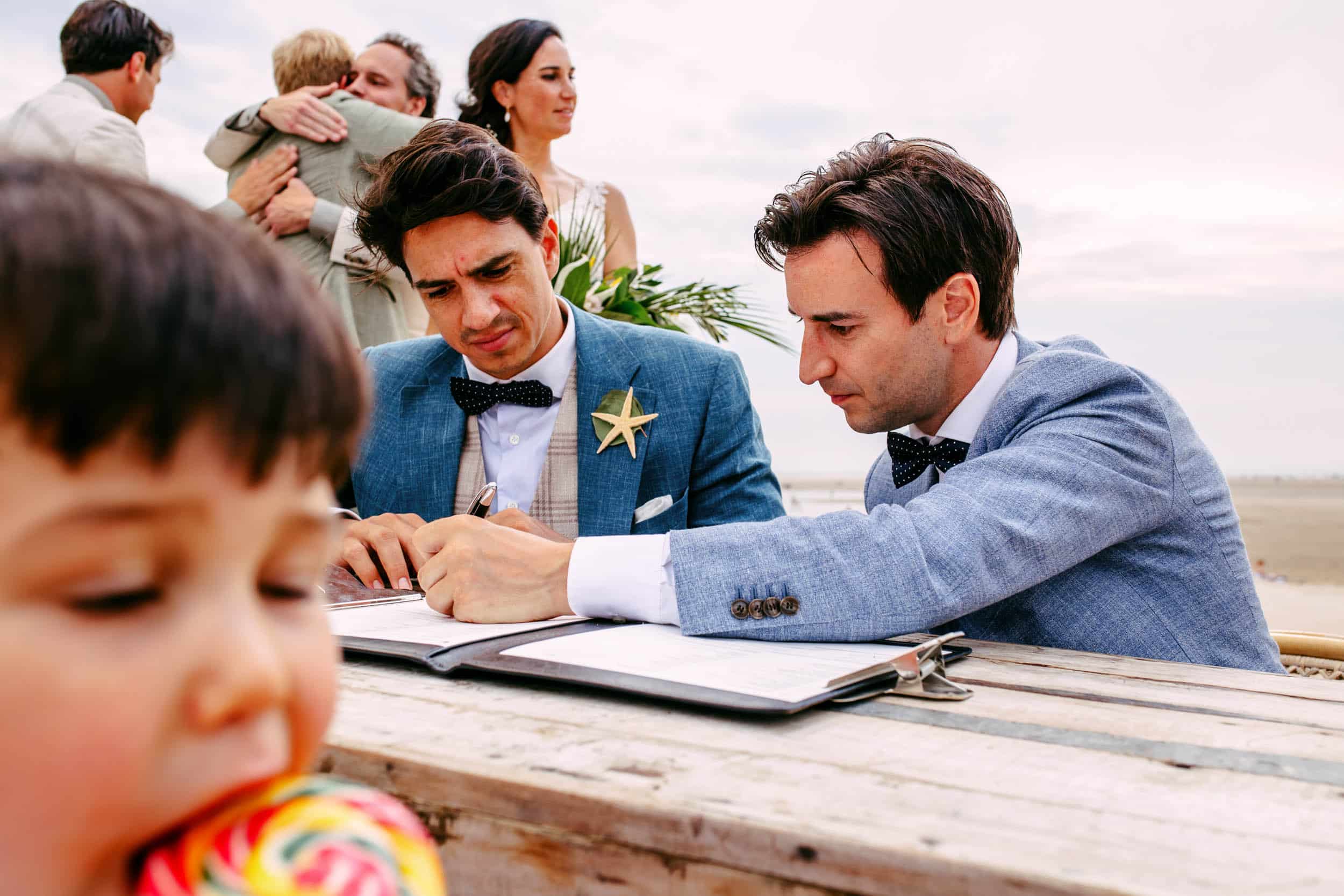 Een man tekent een trouwakte met een jongetje aan tafel en legt het perfecte huwelijksmoment vast door de lens van een trouwfotograaf.