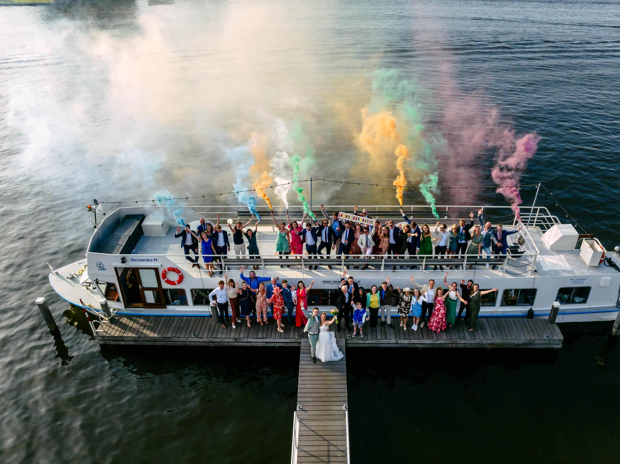 Een groep mensen die op een boot staat tijdens het perfecte huwelijksfeest, waarbij gekleurde rook een prachtig trouwfotograafmoment creëert.
