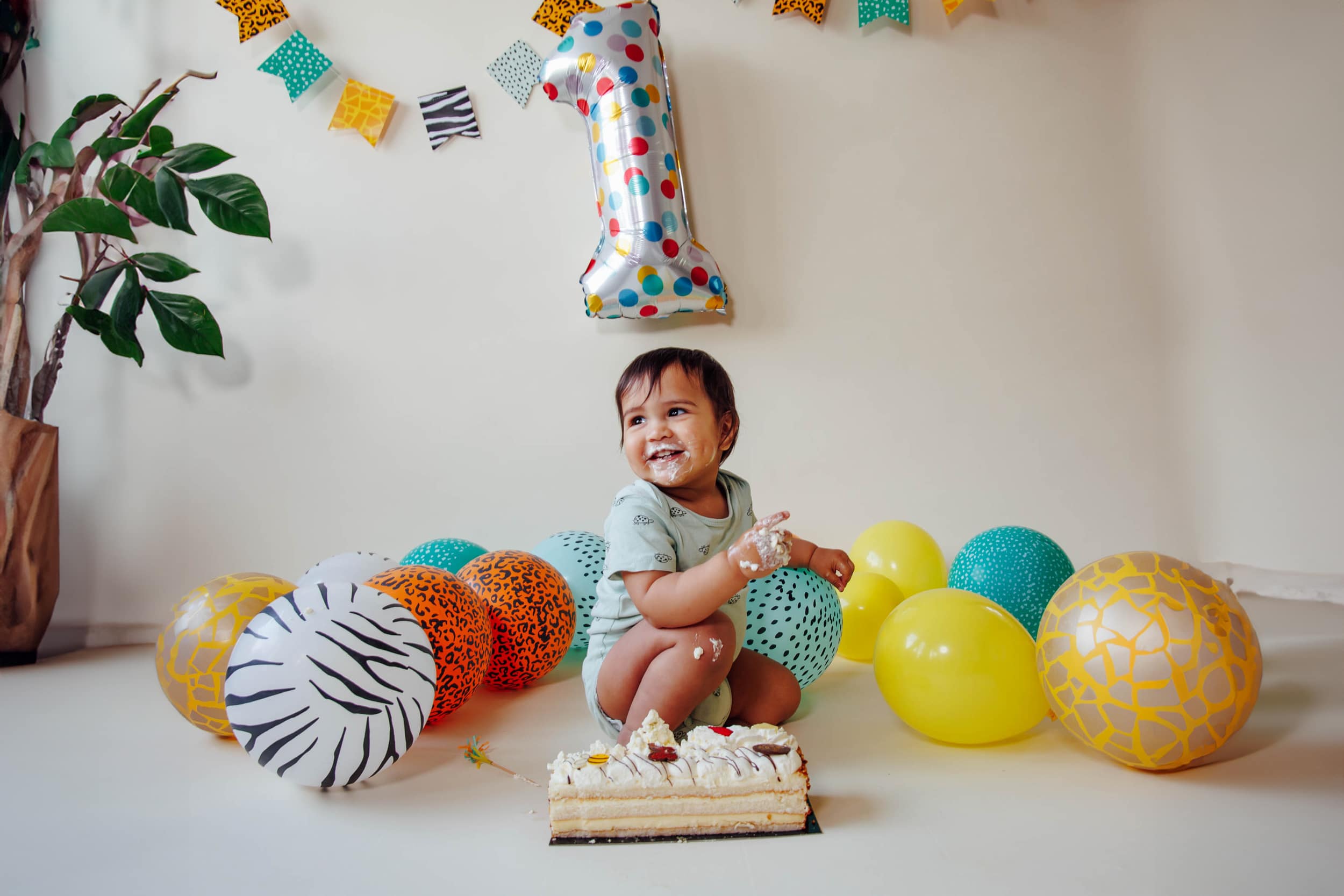 Een peuter zit op de grond met een taart, omringd door kleurrijke ballonnen en een grote '1'-ballon, wat duidt op een eerste verjaardag. Het kind heeft taart op zijn gezicht en handen.