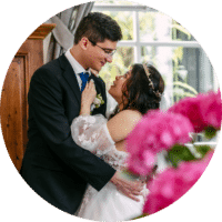 Bruid en bruidegom staren elkaar aan terwijl ze elkaar omhelzen, met roze bloemen op de voorgrond en een raam op de achtergrond.