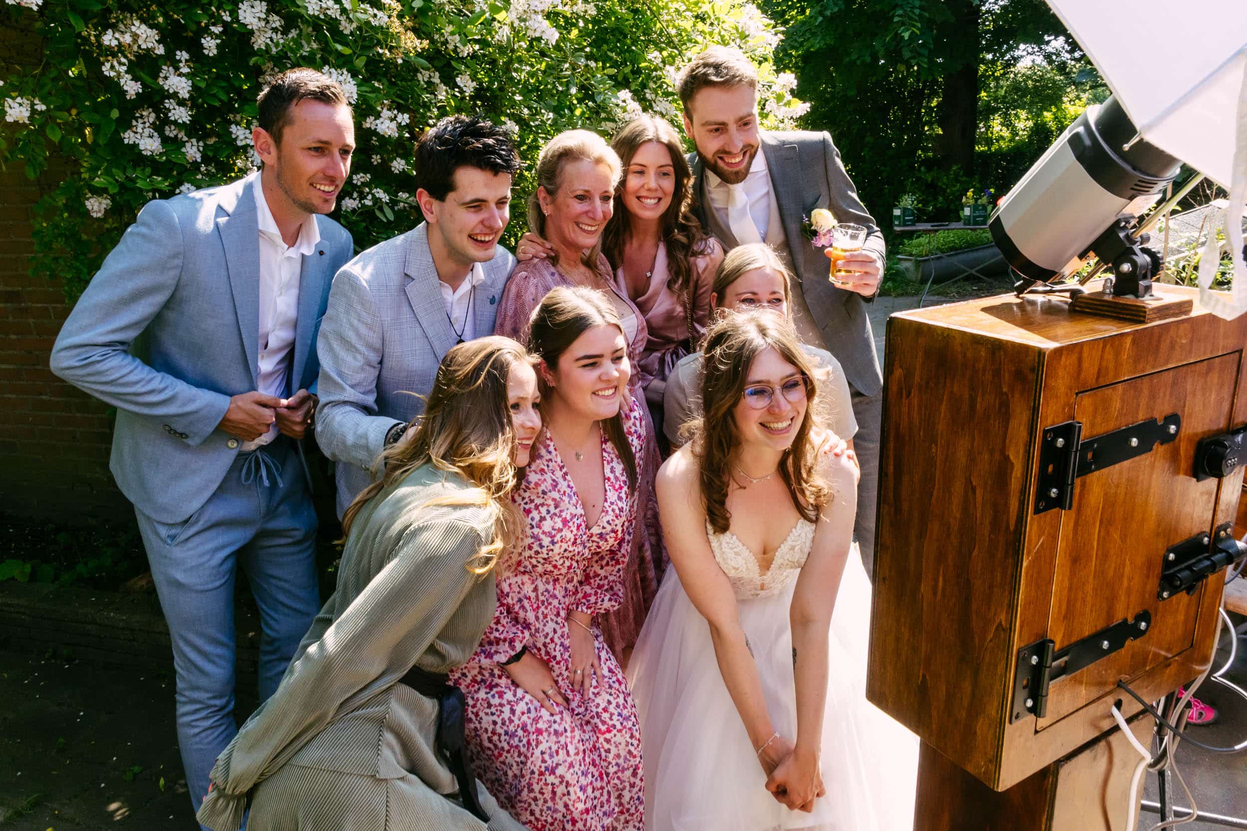 A group of people, dressed in formal and semi-formal attire, gather close together and smile at a vintage-style photo booth outside with greenery in the background.